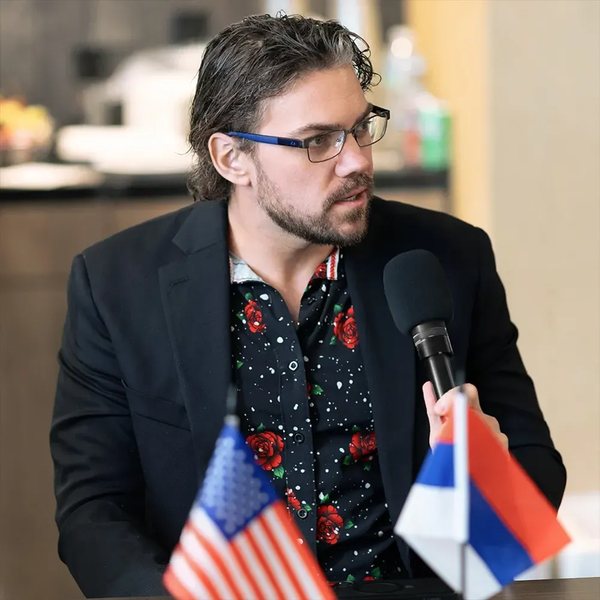 Michael Griswold speaking at a microphone, with an American flag beside him